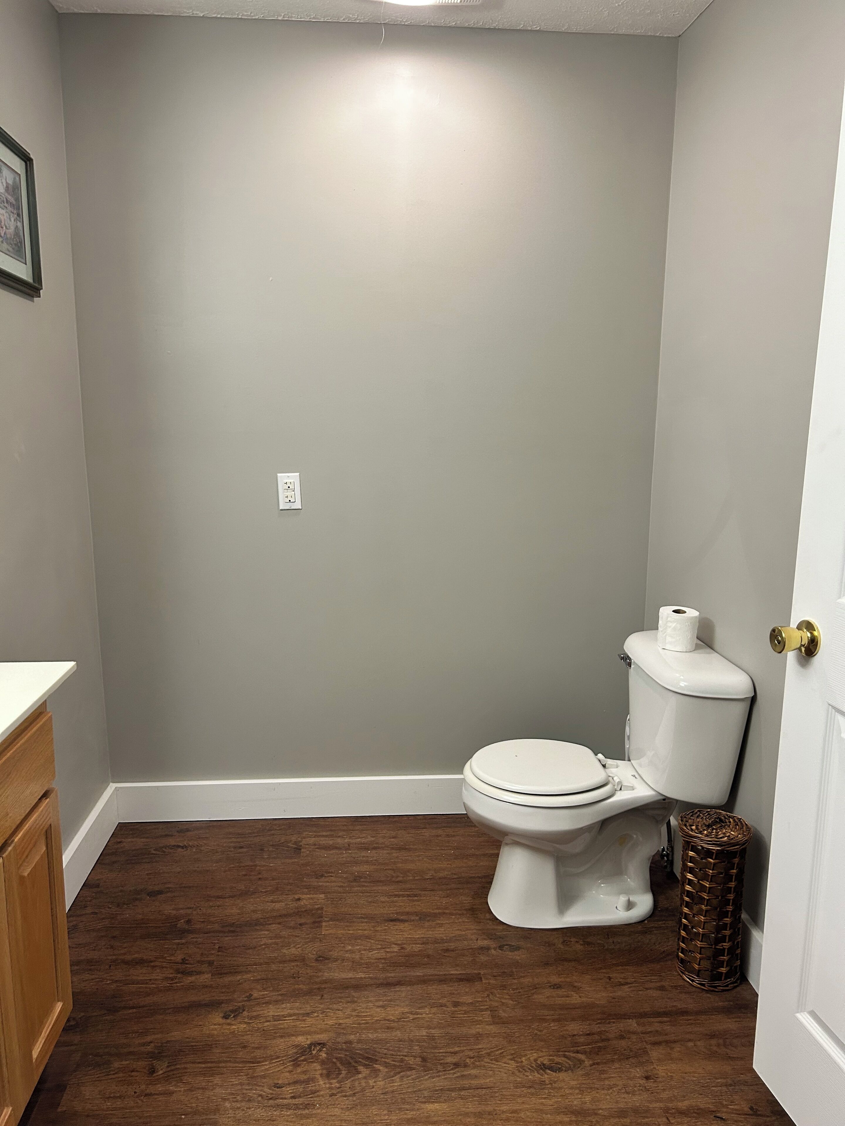 Un baño con inodoro, armarios de madera, una puerta blanca y una cesta.