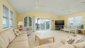 A spacious living room with a sofa, a coffee table, and a television. There are ceiling fans, a window with blinds, and a door leading to a balcony. - 5770- What A View - Oceanfront Getaway Near Jennette's Pier (Nags Head)