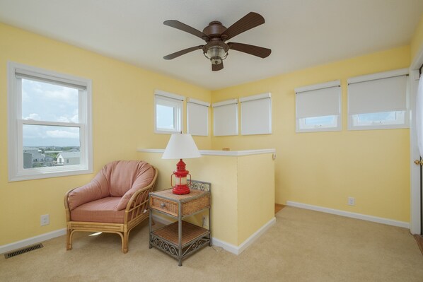 A room with a ceiling fan, a wicker chair, a small table with a red lamp, and a view through a window.