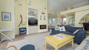 A living room with a blue sofa, a yellow coffee table, and a bookshelf. - 5593-Craven Haven, Nags Head OBX Retreat | Pool & Sunset Views (Nags Head)