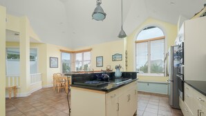 A kitchen with a central island, a dining area with chairs, a refrigerator, and a window with blinds. - 5593-Craven Haven, Nags Head OBX Retreat | Pool & Sunset Views (Nags Head)
