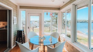 Un espace repas avec une table en verre et des chaises blanches, un plancher en bois et de grandes fenêtres donnant sur l’eau.