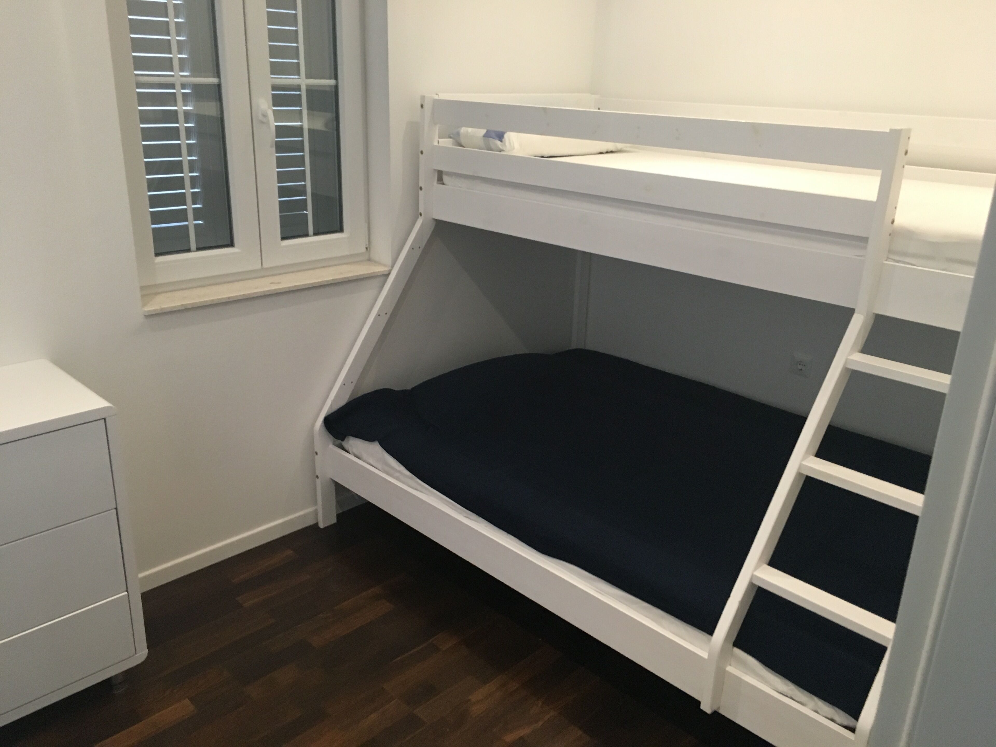 A bunk bed with a ladder, a small bedside table, and a window with blinds.