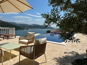An infinity pool with a view of a lake and mountains, surrounded by wicker furniture and a stone deck.
