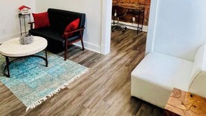 Una sala de estar con un sofá negro, un sillón blanco y una mesita de centro. Hay un cuadro en la pared y una pared de ladrillo al fondo.