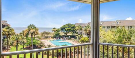 A balcony with a view of a swimming pool, palm trees, and a beach in the background.