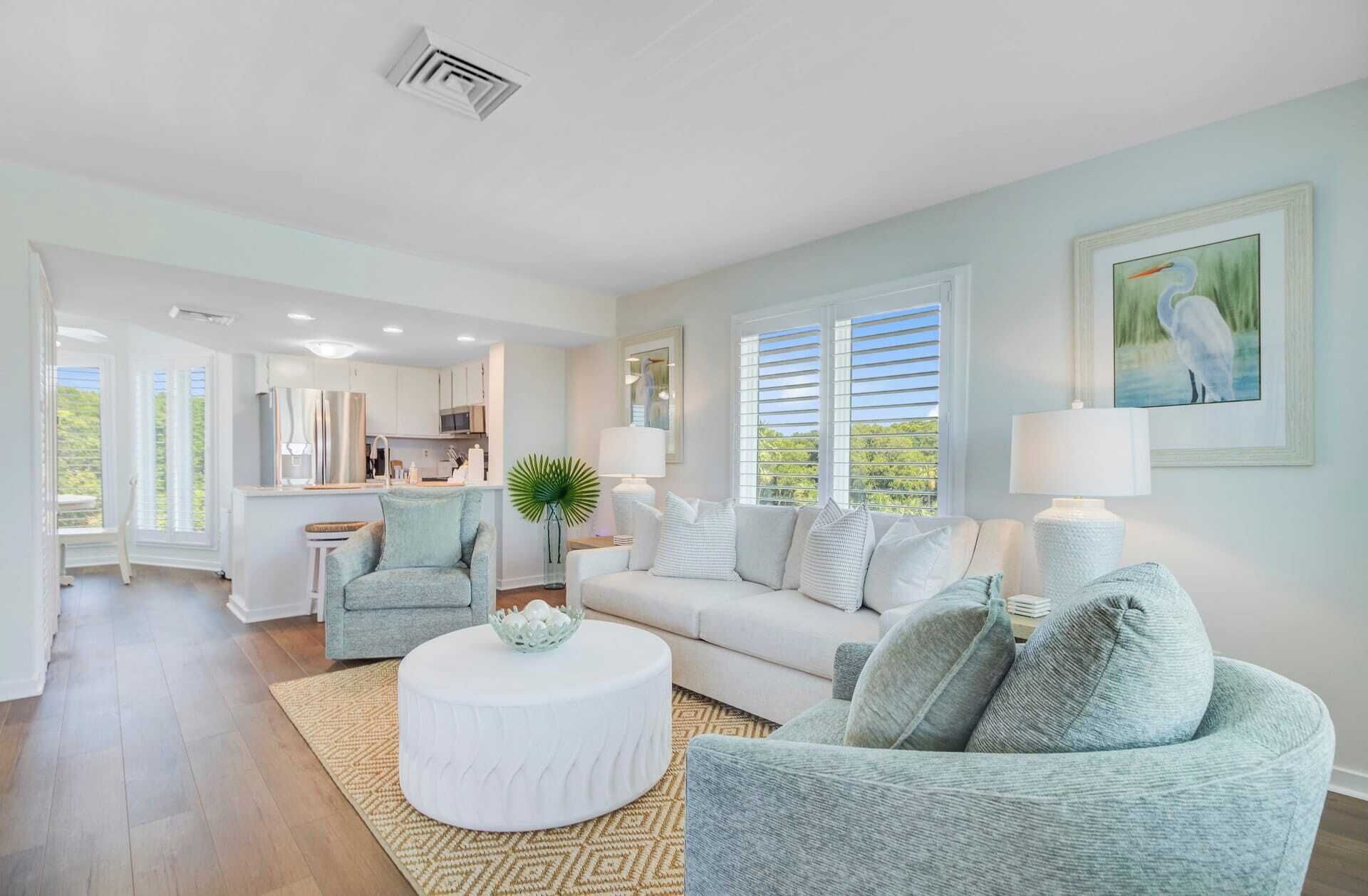 A spacious living room with a white sofa, a round coffee table, and a light blue armchair. There is a kitchen in the background with white cabinets and a dining area.