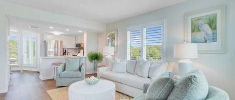 A spacious living room with a white sofa, a round coffee table, and a light blue armchair. There is a kitchen in the background with white cabinets and a dining area.