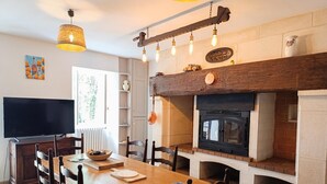 A dining area with a wooden table, chairs, a television, and a fireplace with a stone mantle. - Gîtes de France® - Le Clo (SAINT MICHEL DE BANNIERES)