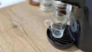 Una cafetera sobre una superficie de madera con jarra de vidrio transparente y una taza pequeña.