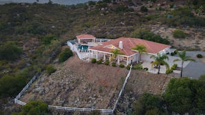 A two-story house with a red-tiled roof, surrounded by a white fence, and located on a hillside with sparse vegetation. - Pool, Views, Hot Tub, Fire Pit & Arcade Vista Clara by Avantstay (Temecula)