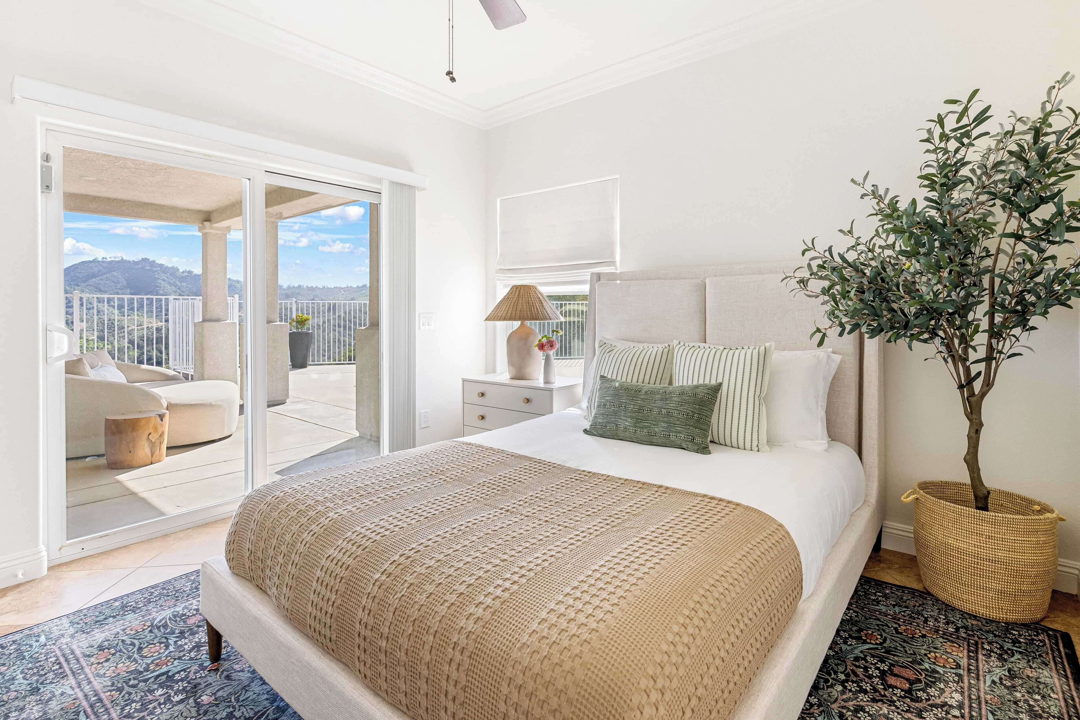 A bedroom with a large bed, a nightstand, a lamp, a potted plant, and a view of a balcony with a sofa and a table.