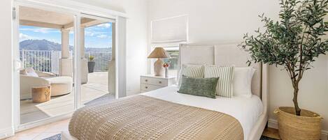 A bedroom with a large bed, a nightstand, a lamp, a potted plant, and a view of a balcony with a sofa and a table.