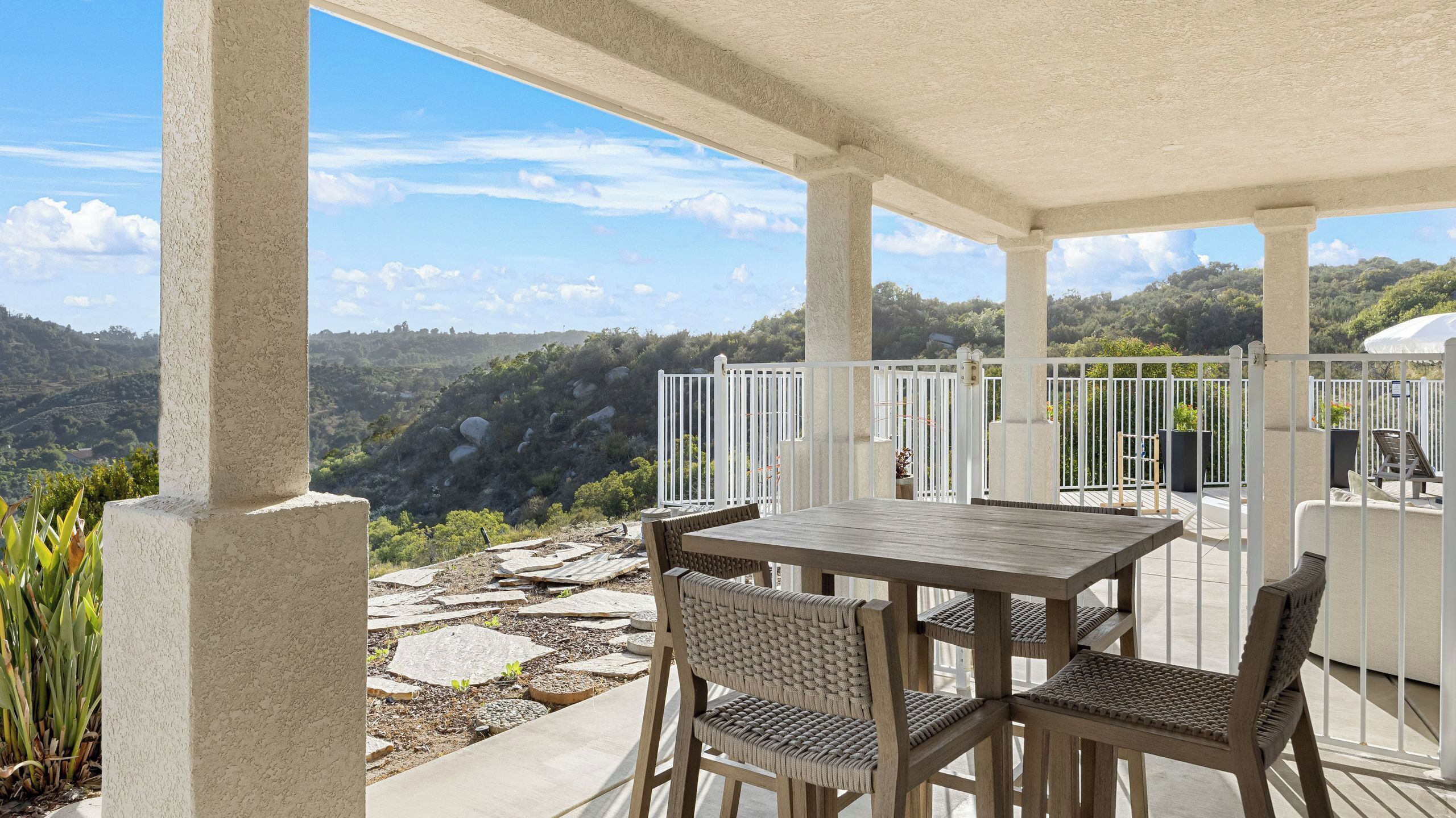 A covered patio with a table and chairs, white railings, and a view of a mountainous landscape.