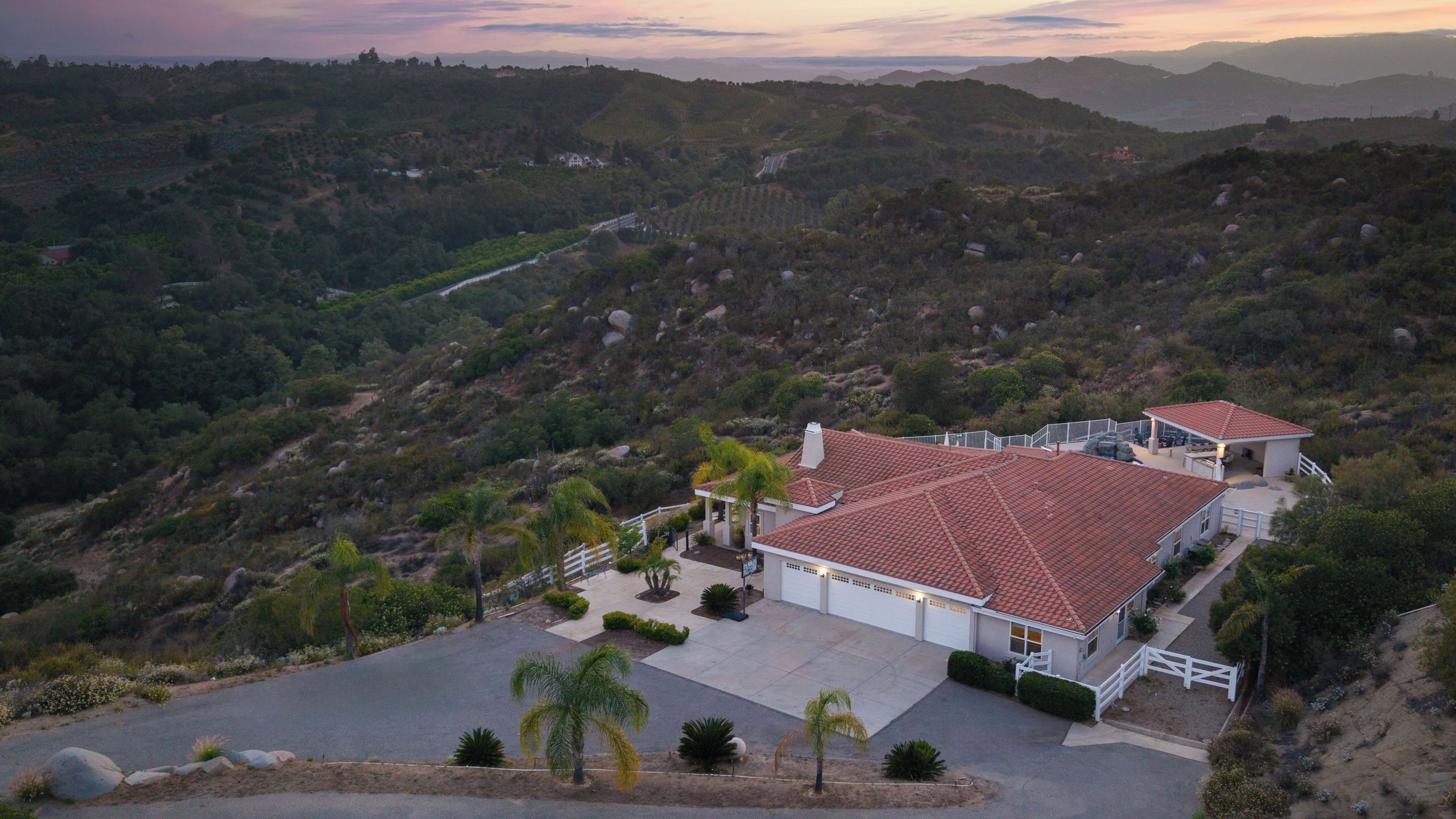 A house with a red-tiled roof, surrounded by a landscaped garden with palm trees, set against a mountainous backdrop.