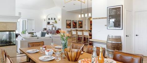 Un coin repas avec une table en bois, des chaises et un lustre. Au fond, on aperçoit un bar avec une enseigne indiquant « BAR ».
