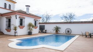 A swimming pool with a tiled deck, a patio area with chairs and tables, and a building with a tiled roof and white walls.
