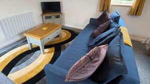 Een moderne woonkamer met een blauwe bank, een houten salontafel, een tv op een standaard en een raam met gordijnen.