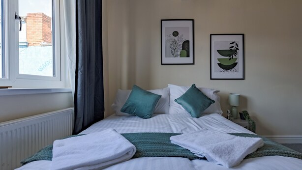 A neatly made bed with white and teal bedding, two framed artworks on the wall, and a window with curtains. - Modern Cozy 2BR- Private Parking- Near WBA (Birmingham)