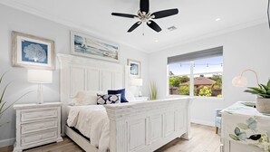 A bedroom with a white bed, a nightstand, a dresser, a ceiling fan, and a large window with a view of greenery. - Casa Blu - Bonita Springs, FL (Bonita Springs)