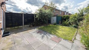 A residential backyard with a paved patio, a grassy lawn, a wooden fence, and a green shed.