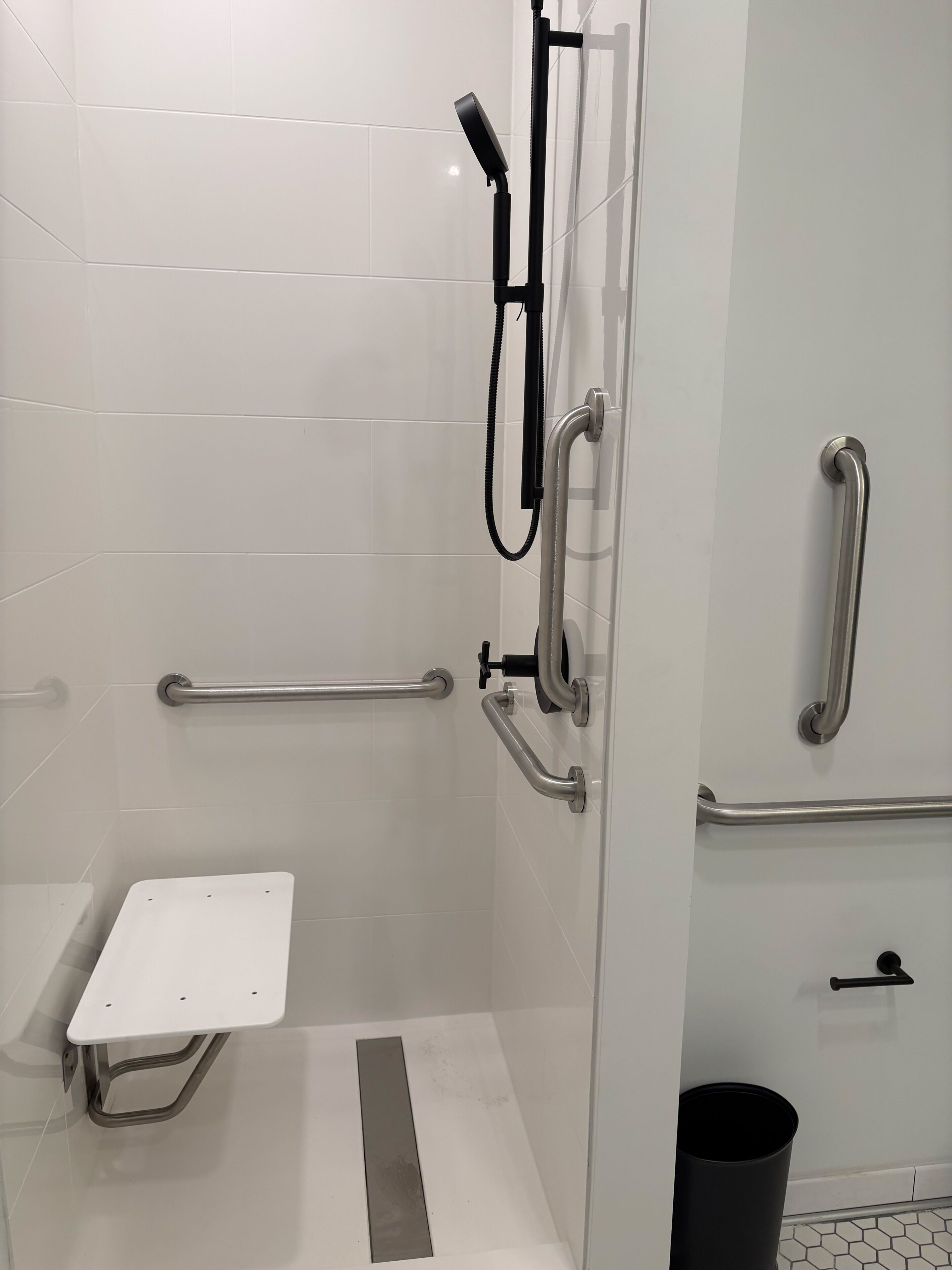 Ein modernes Badezimmer mit weißen Fliesen, einer Dusche inklusive Duschbrause, Haltegriffen und einer weißen Sitzbank.