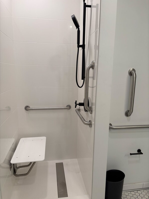A modern bathroom with white tiles, a shower with a handheld showerhead, grab bars, and a white bench. - The Lostro – a boutique hotel (Athens)
