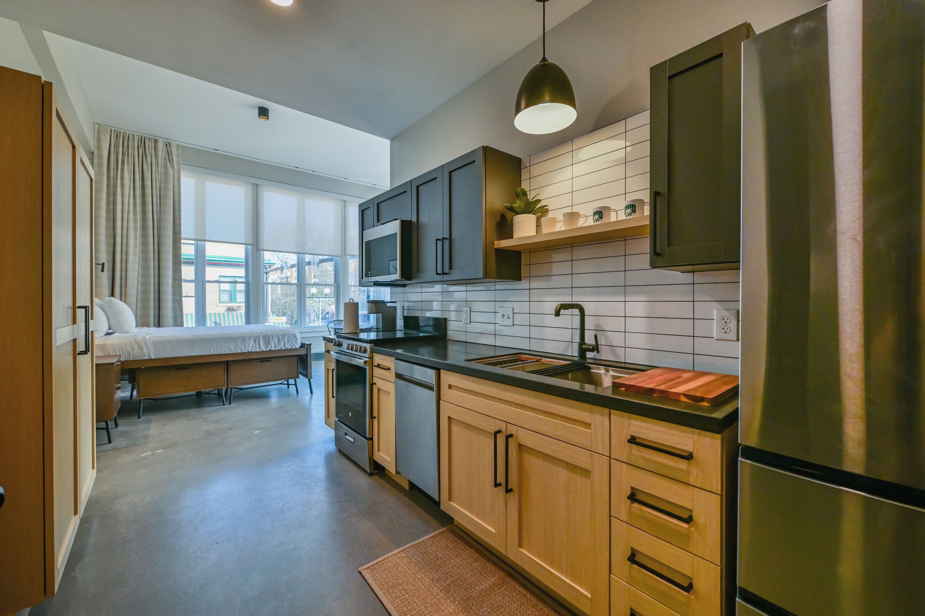 A compact kitchen with a microwave, stove, and sink, a small dining area with a bench, and a large window providing a view of the city.