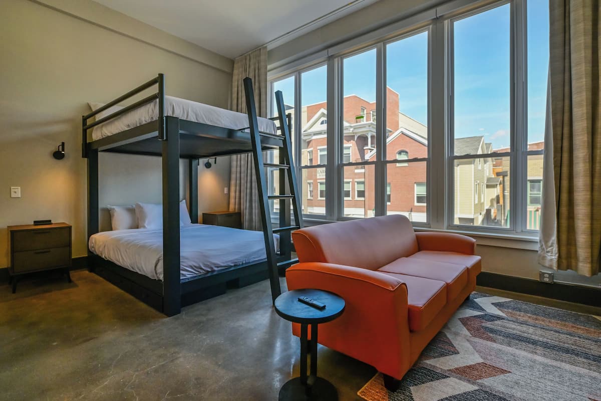 A room with bunk beds, a sofa, a small table, and large windows with a view of buildings.