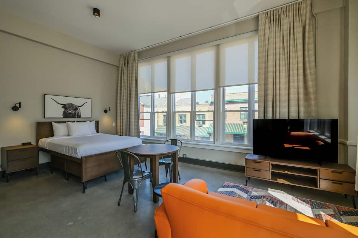 A hotel room with a bed, a TV, a sofa, a dining table, and a window with curtains.