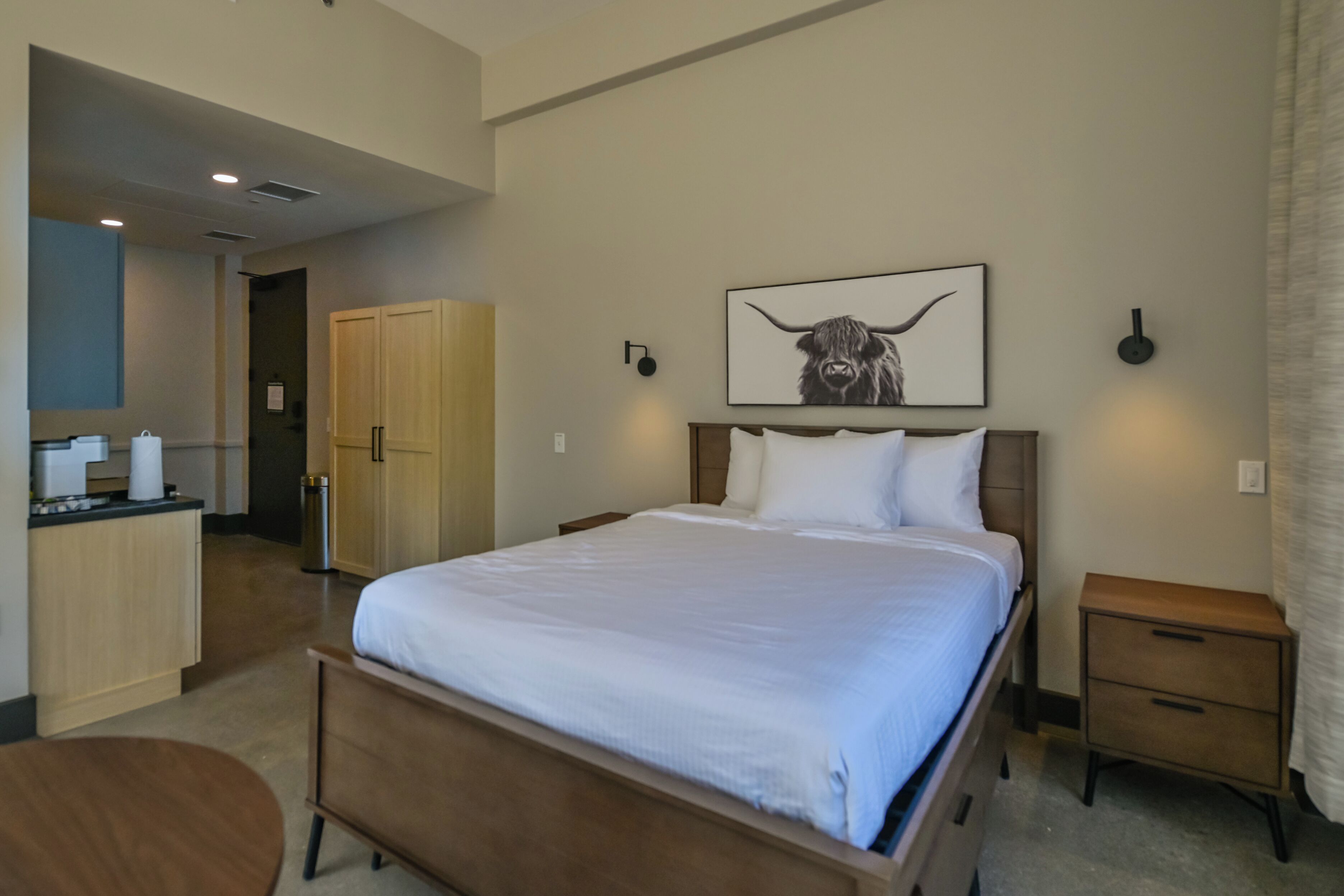 A hotel room with a bed, bedside table, a small desk with a chair, a wardrobe, and a picture of a bull on the wall.