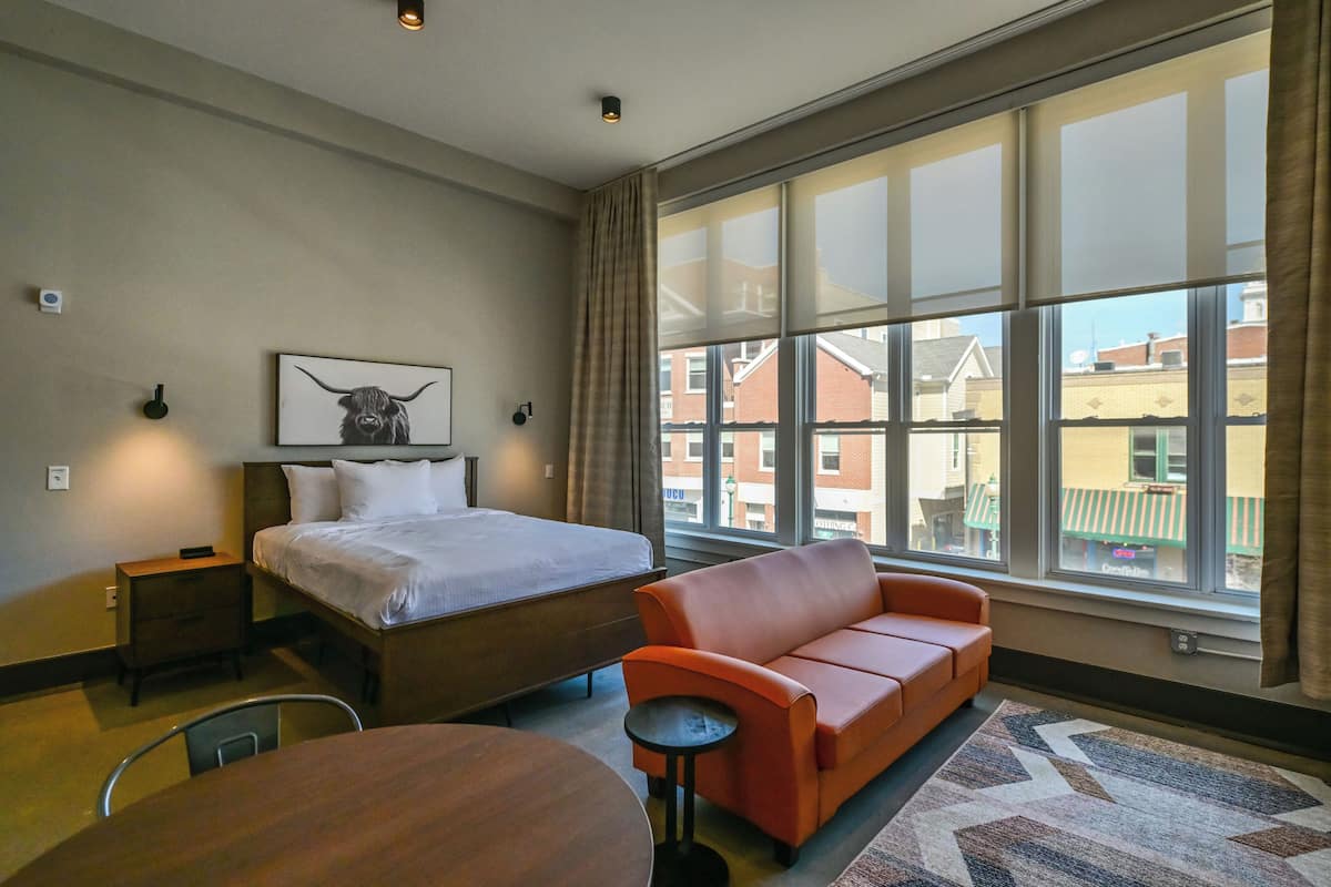 A hotel room with a bed, a sofa, a dining table, and large windows.