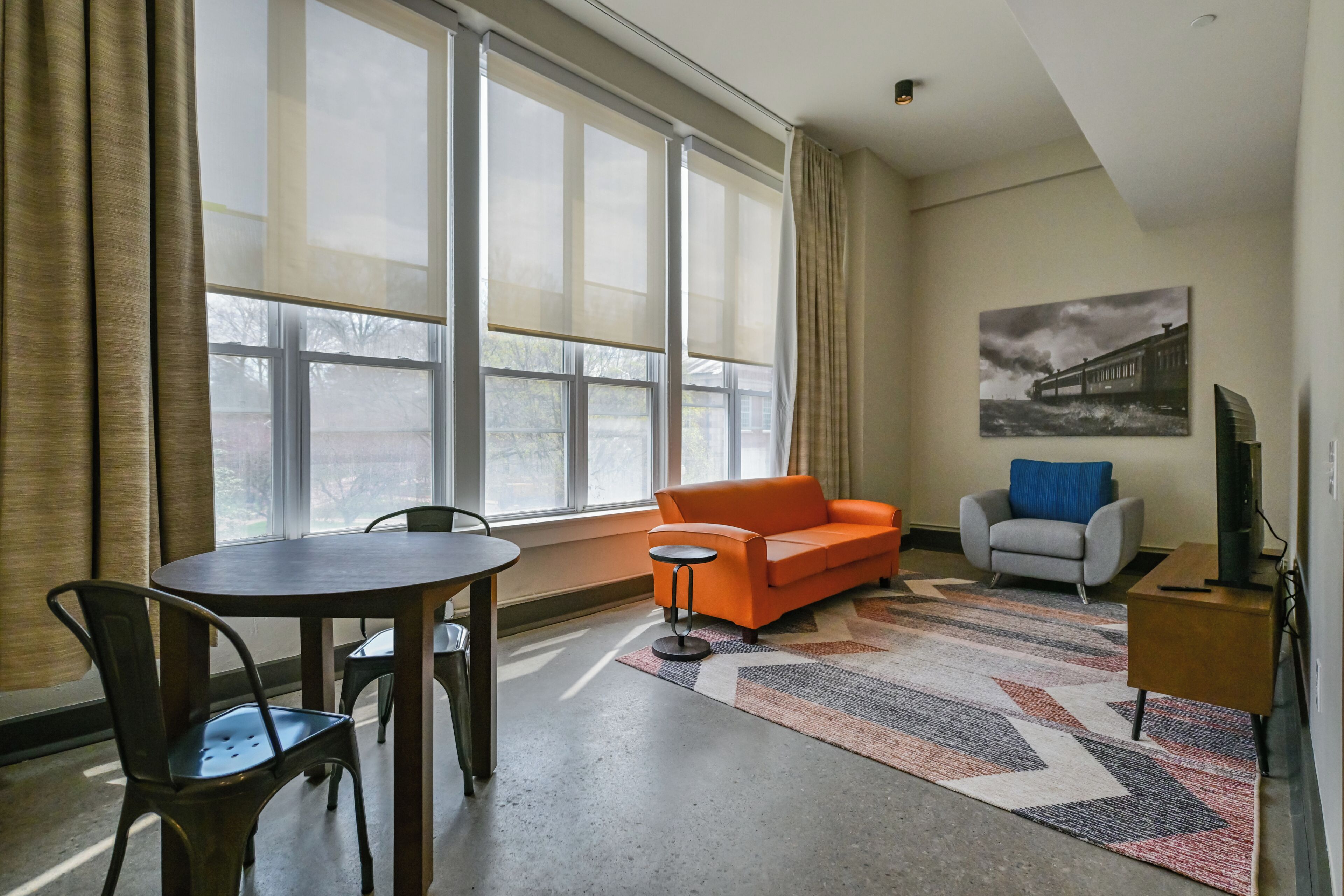 A room with a dining table, chairs, an orange sofa, a gray armchair, a rug, and a painting on the wall.
