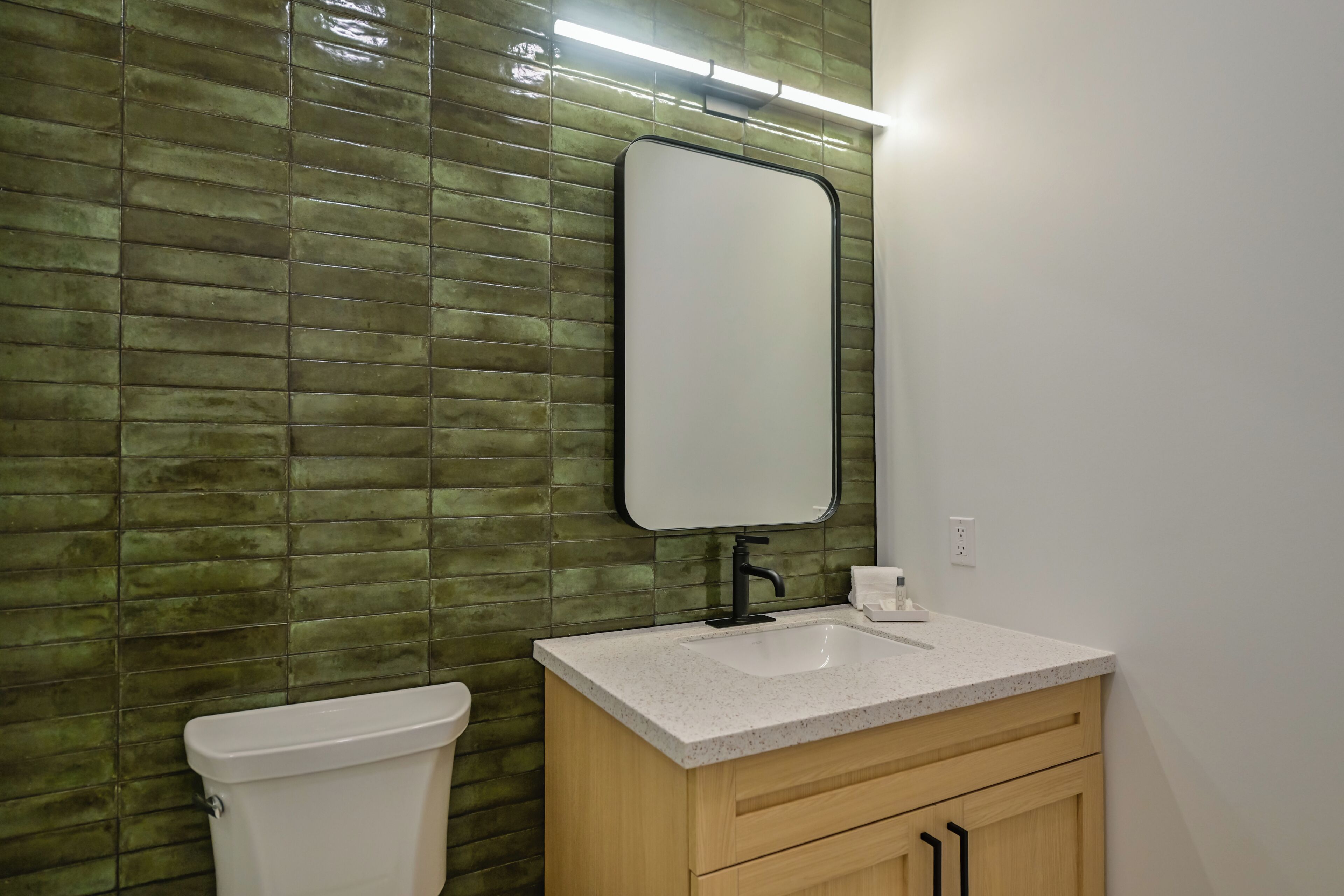 Une salle de bain avec un mur recouvert de carreaux verts, des toilettes blanches, un lavabo avec un miroir et des meubles en bois.