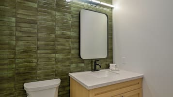 Une salle de bain avec un mur recouvert de carreaux verts, des toilettes blanches, un lavabo avec un miroir et des meubles en bois.