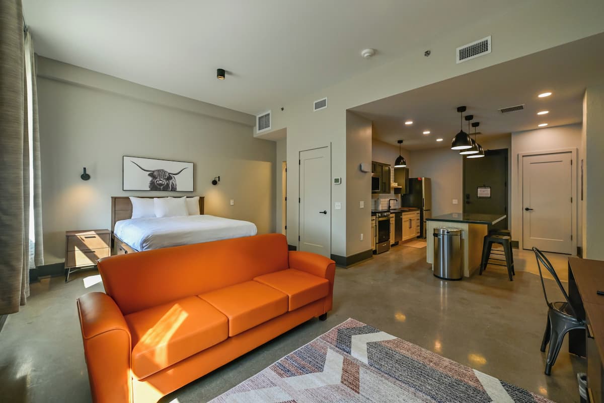 A modern hotel room with a bed, orange sofa, kitchenette, and a dining area.