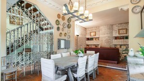 A dining area with a large table, white chairs, a chandelier, and a brick wall with shelves.