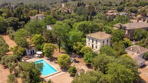 An aerial view of a property with a swimming pool surrounded by trees, a large house with a tiled roof, and a hilly landscape in the background.