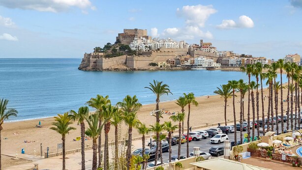 Una ciudad costera con una fortificación histórica, una playa de arena bordeada de palmeras y un estacionamiento con coches.