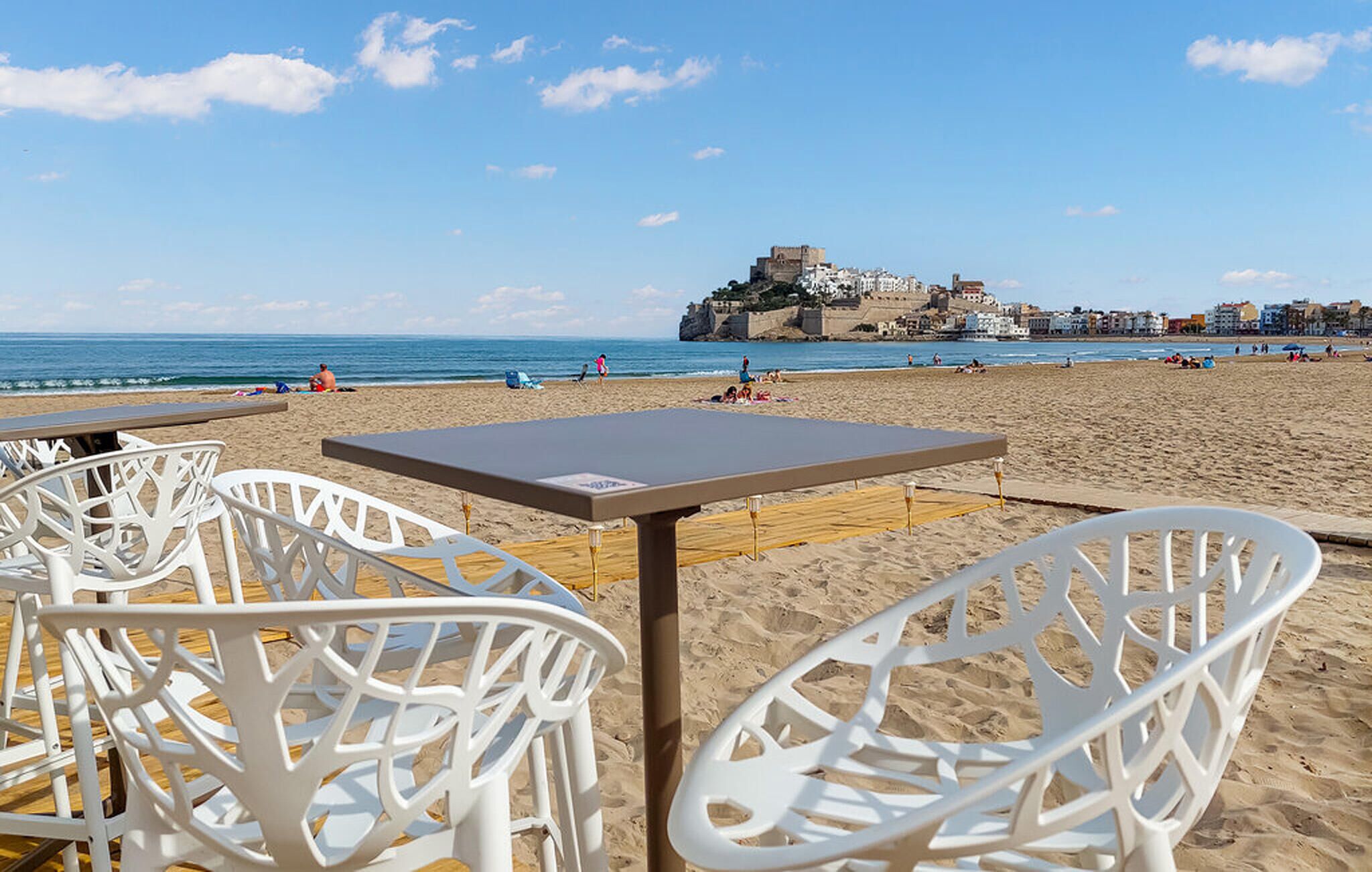 A beach with white plastic chairs and a table, a castle-like structure in the background, and the sea.