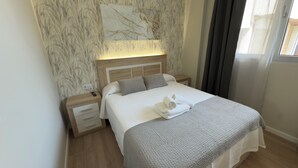 Una cama bien hecha con una manta gris y una toalla enrollada, una mesita de noche de madera con un control remoto, y una pared con diseño tipo mármol.