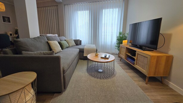 A modern living room with a sectional sofa, a round coffee table, a TV on a wooden stand, and a large window with sheer curtains.