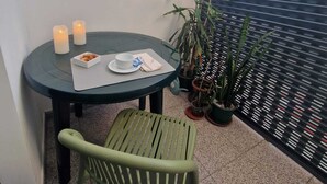 A small round table with a chair, candles, a plate with food, and a cup. There are potted plants and a window with blinds in the background.