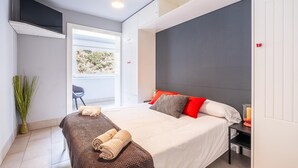 A modern bedroom with a bed, a chair, a small table, and a window with a view of greenery.