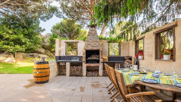 An outdoor dining area with a stone barbecue, wooden chairs, and a table set for a meal.