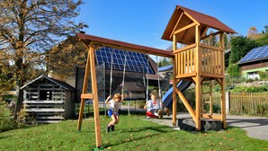 A playground with swings and a slide, surrounded by trees and a house with solar panels. - Apartment 'Wiesenblick' with Shared Terrace, Shared Garden and Wi-Fi (Schramberg)