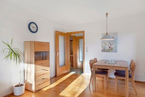 A room with a wooden cabinet, a dining table with chairs, a potted plant, a clock on the wall, and a door leading to another room.