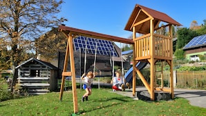 A playground with swings and a slide, surrounded by trees and a house with solar panels. - Apartment 'Gartenblick' with Shared Terrace, Shared Garden and Wi-Fi (Schramberg)