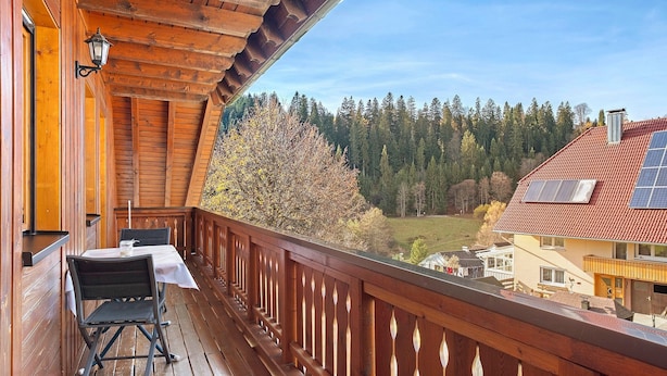 A wooden balcony with a table and chairs, overlooking a forested area and a house with solar panels. - Apartment 'Gartenblick' with Shared Terrace, Shared Garden and Wi-Fi (Schramberg)