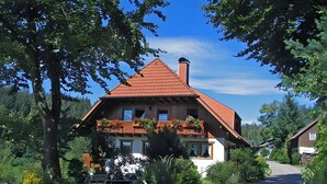 A two-story house with a red roof, a balcony with plants, and a driveway leading to the house. - Apartment 'Gartenblick' with Shared Terrace, Shared Garden and Wi-Fi (Schramberg)